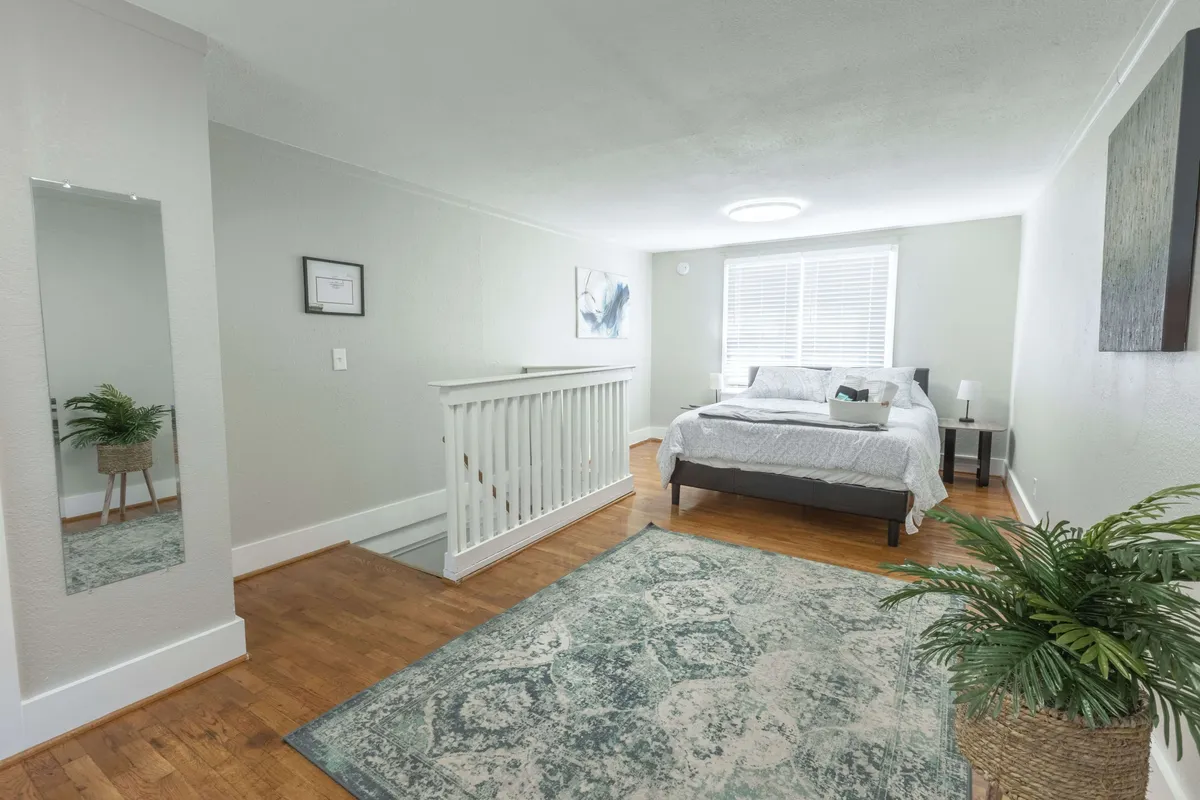 Featuring a bright bedroom with a bed, rug, mirror, potted plant, and artwork, this image is filled with natural light and hints at the start or end of a day. The inviting atmosphere suggests relaxation after a day out or restful preparation before heading into the city.