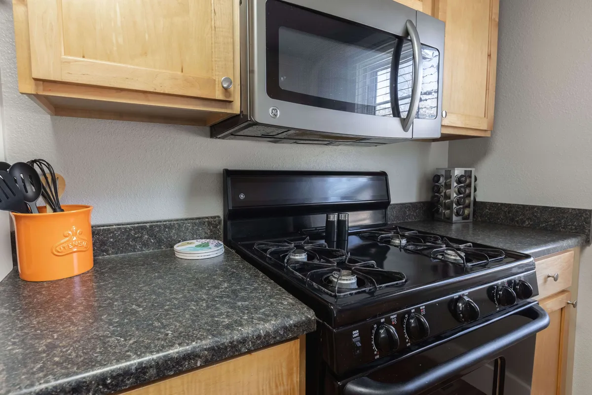 The photograph highlights a section of the kitchen, focusing on a stainless steel microwave, sleek black gas stove, attractive wooden cabinetry, and a vibrant orange utensil holder. The overall look is both inviting and practical, suggesting the kitchen is ready for any culinary adventure.