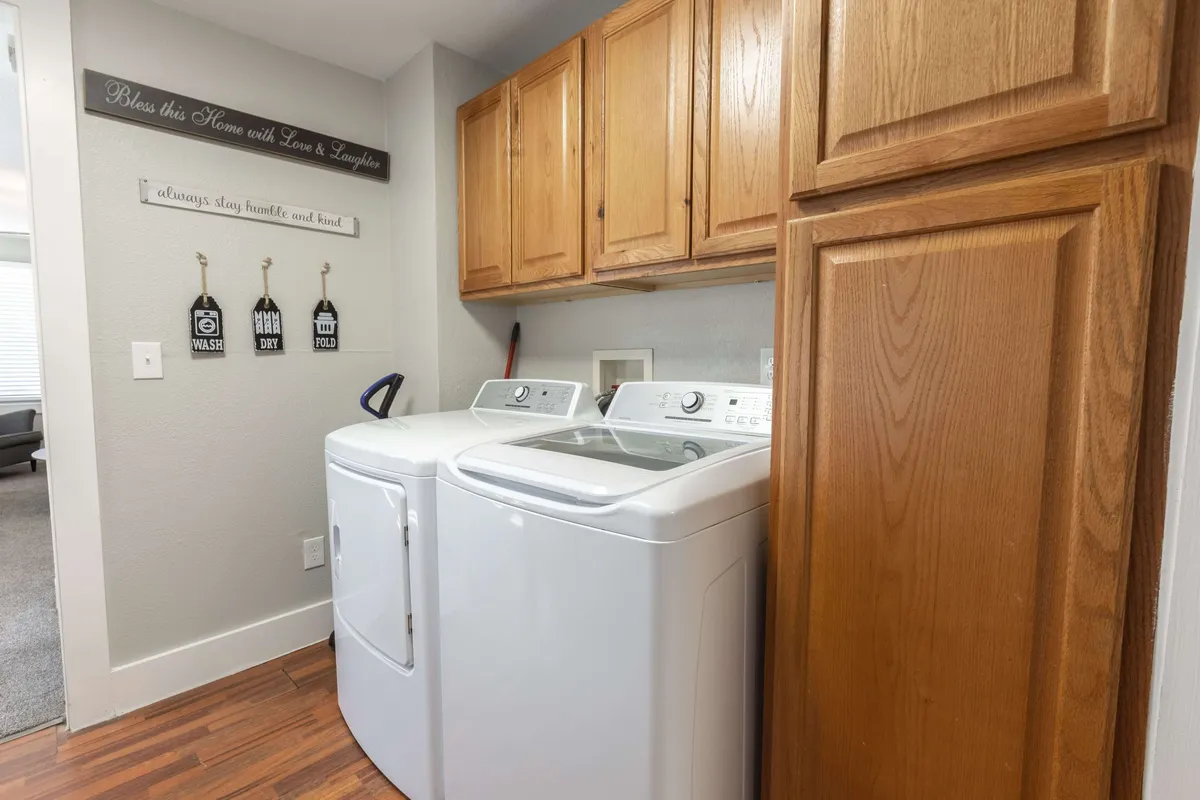 This image depicts a well-organized laundry area featuring a washing machine and dryer beneath wooden cabinetry, decorated with homey signs and practical touches. The space feels inviting and functional, suggesting opportunities for casual conversation while handling simple chores.