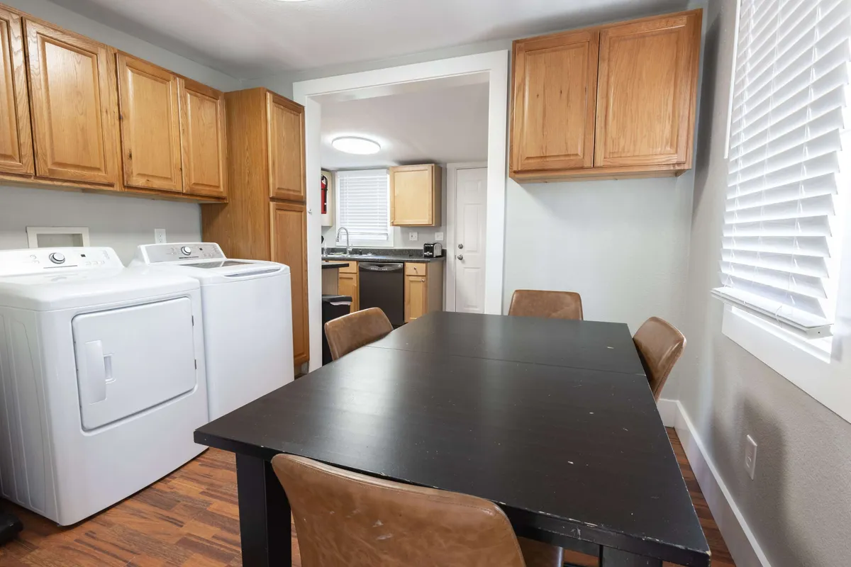 A view of a dining table set in a bright, homey space with cabinetry and washing machines visible in the background. The room has a welcoming, lived-in feel, suggesting it’s a hub for family meals and daily routines.