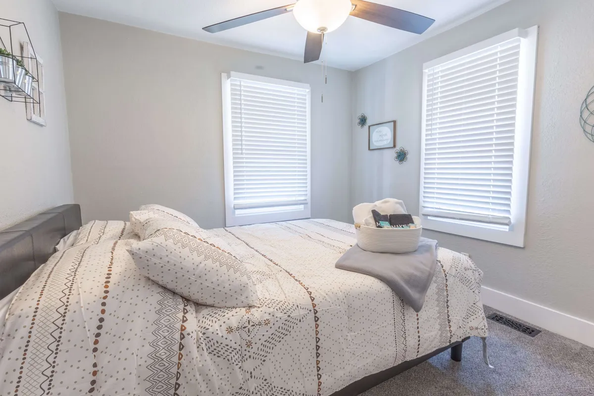 A tranquil bedroom scene with a comfortable bed, large windows letting in warm sunlight, decorative wall art, and a ceiling fan above. The atmosphere encourages relaxation, napping, or quiet moments spent reading.