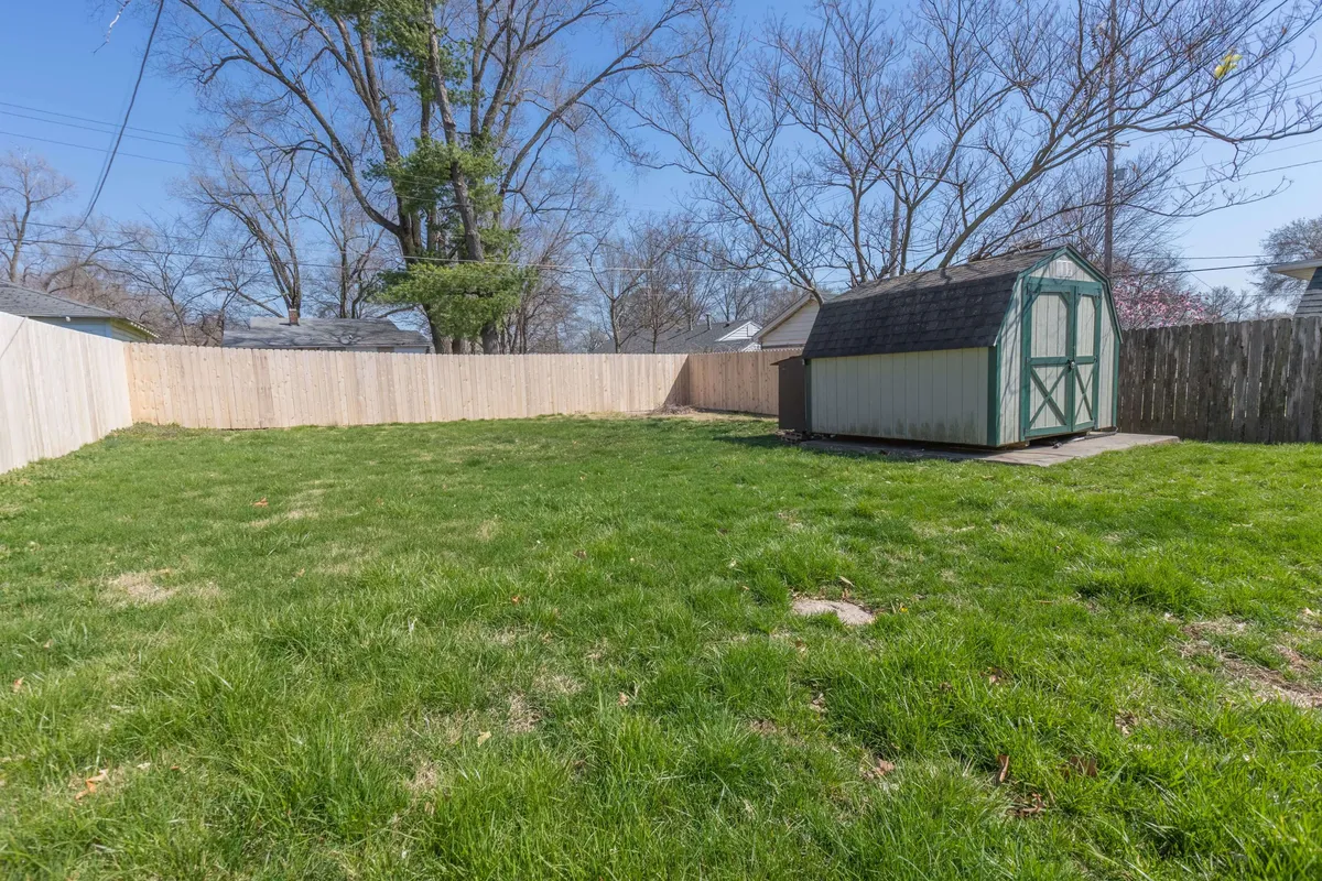 This image presents an expansive green lawn bordered by mature trees, with a storage shed off to one side. The open, sunny yard provides ample space for outdoor activities such as gardening, family games, or relaxing evenings. The scene conveys a sense of privacy and tranquility, with natural elements and open sky.